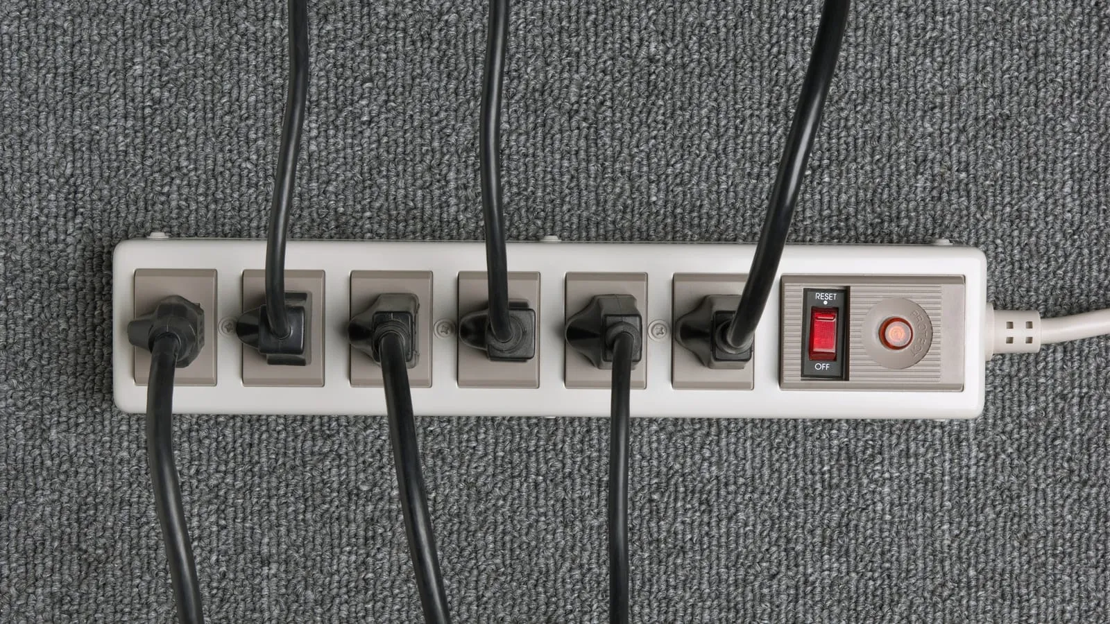 A six-outlet power strip on a grey carpet with a plug in every socket