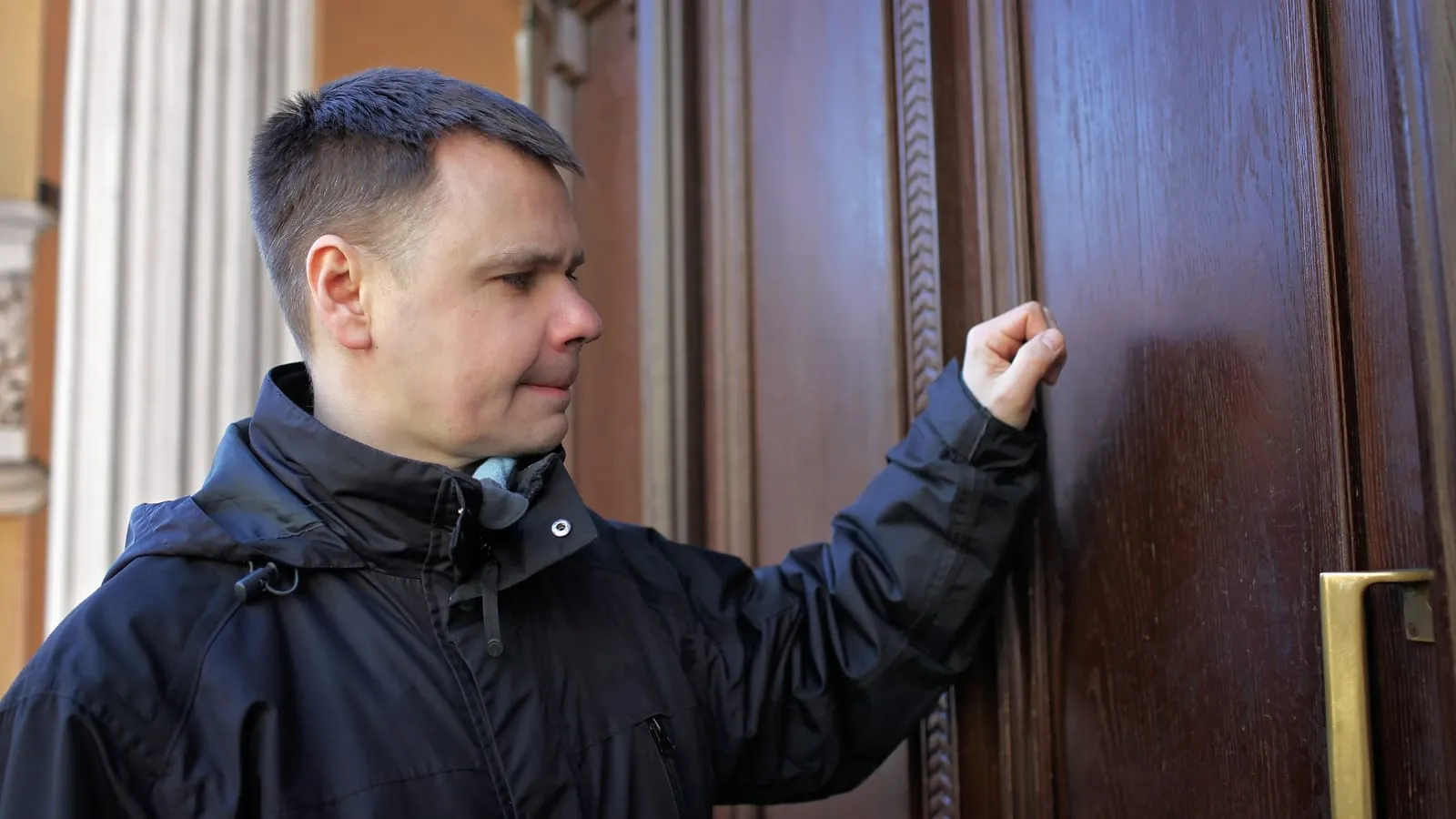 A man in a dark jacket about to knock on a tall dark wooden door, his fist raised next to a brass handle